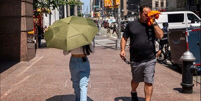 stock image from Getty. Man wipes sweat from his face as he walks along a city street and crosses paths with a woman holding an umbrella to block the sun.