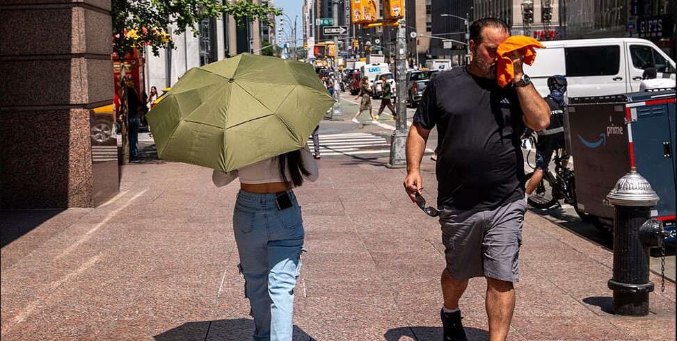 stock image from Getty. Man wipes sweat from his face as he walks along a city street and crosses paths with a woman holding an umbrella to block the sun.