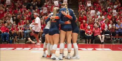 Image of Illini volleyball players huddling on court, this time at Wisconsin so the crowd is all wearing red.