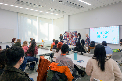Image depicts individuals looking toward the front of a classroom while three people stand at the front, two of which are holding a quilt.