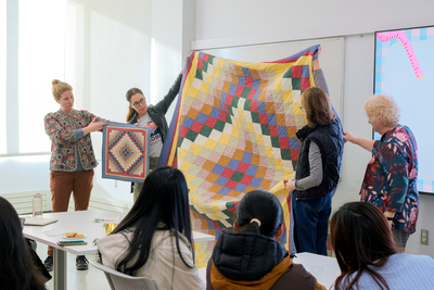 Kathryn Burden (standing on the far left) holds up a small quilt, while two volunteers (center) hold up the larger version of the same quilt during a Jan. 7 workshop inside the Siebel Center for Design. Jennie, Kathryn's mom, stands on the far right and explains the story behind the quilt.