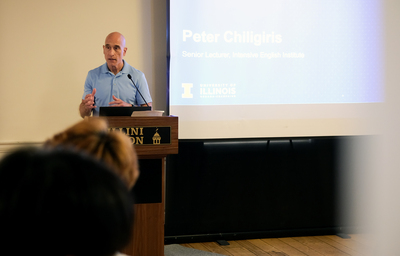 Peter Chiligiris speaks at a podium during a program ceremony as a presentation slide bearing his name is displayed on a screen behind him.