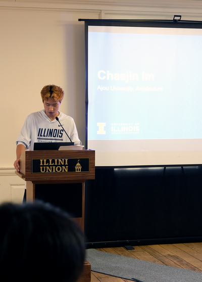 Chaejin Im stands at a podium in the Illini Union and delivers a speech during a ceremony. A presentation slide displaying his name and Ajou University affiliation is projected on a screen behind him.