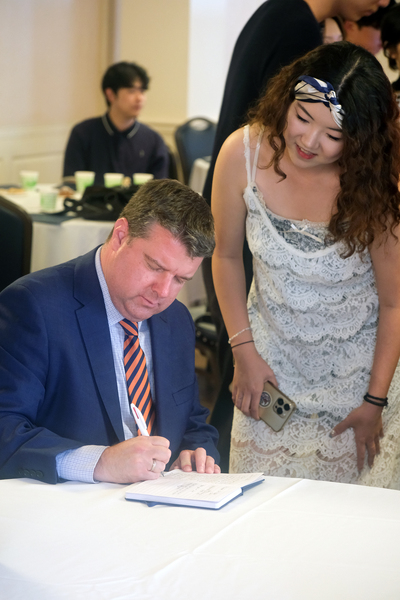 Jihyun “Tiffany” Kim stands beside Matt Rosenstein as he writes a message in a notebook during a gathering at the Illini Union. Other attendees are visible in the background.