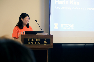 Harin Kim stands at a podium in the Illini Union and delivers a speech during a ceremony. A presentation slide displaying her name and Ajou University affiliation is projected on a screen behind her.