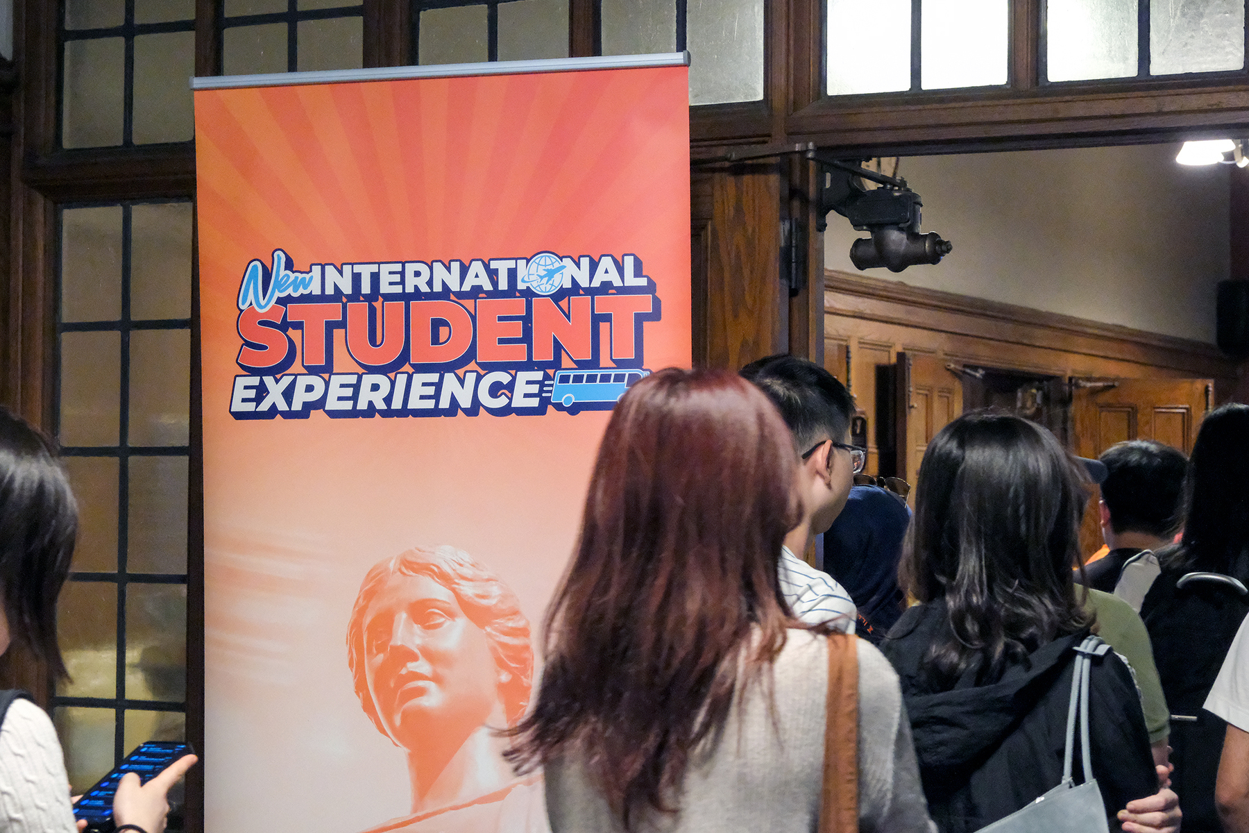 A group of students stands in line inside a wood-paneled hall beside a tall orange banner that reads “New International Student Experience.” The banner features a stylized campus statue and bright sunburst design, while large windows and architectural details are visible in the background.