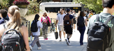Students walk on the University of Illinois Urbana-Champaign campus as a bus drives by in the background.