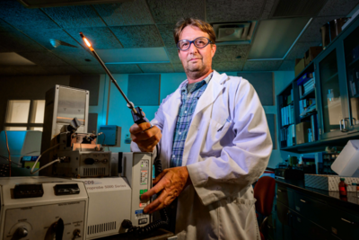 A man wearing goggles and a lab coat holding a device.