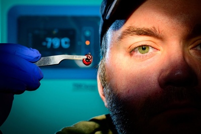 Man holds up a tick with tweezers, only one eye of his face lit.