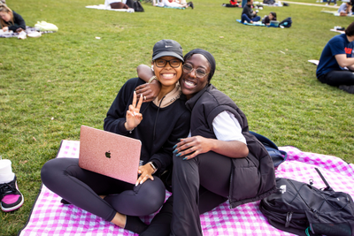 Two students smiling while sitting on a blanket in a green space