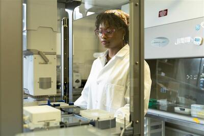A Black woman stands in a lab and looks through a glass case. She is wearing a white lab coat.