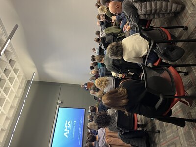 A photo taken from the back of the room full of people sitting in chairs and a man standing behind a podium at the front of the room.
