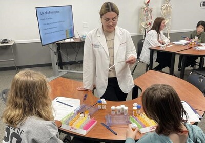 A pharmacist speaking to two youngers students who are seated at a table with pill bottles and papers on it