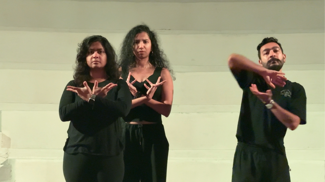 Move. Three dancers in black stand under soft lighting, hands posed in expressive gestures, facing the camera against a pale wall.