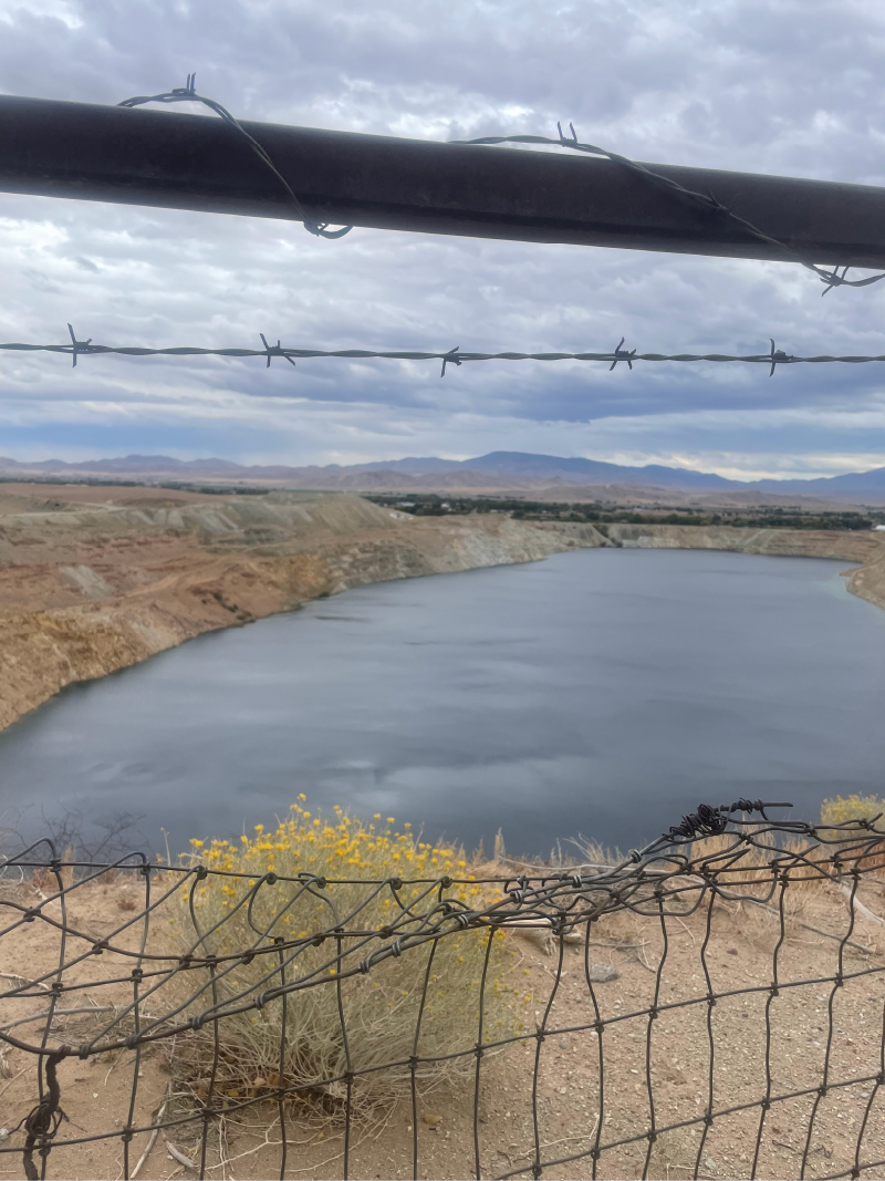 Coppered Water and Barbed-Wire Sagebrush in the Desert Wasteland. Water fills an open mine pit with mountains in the back in a desert landscape, with sagebrush all amidst barbed wire fencing.
