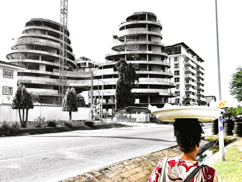 Phases and Faces of Urban Spaces. Three multi-story buildings to the left and a female street hawker to the right separated by the street.