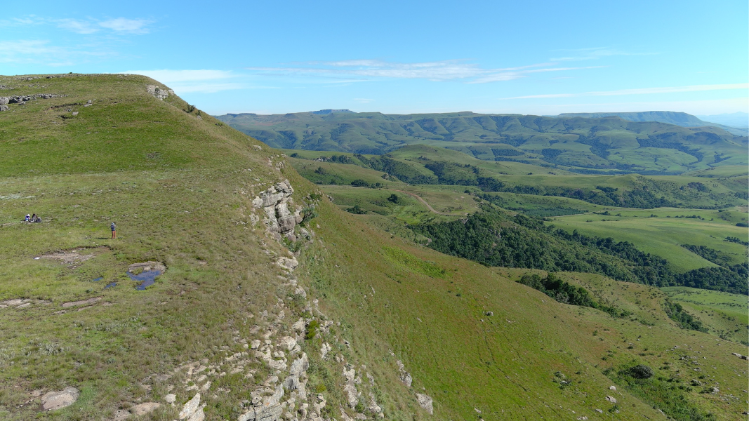 Finding Resilience in Unlikely Places. The shoulder of a mountain with flat bare patches, three small figures, and a landscape of green rolling hills.